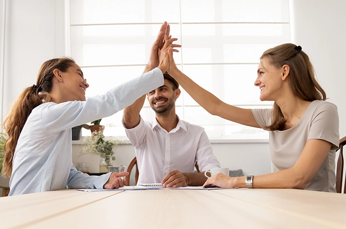 Zwei Frauen und ein Mann geben sich ein gemeinsames High Five, während sie am Tisch in einer Besprechung sitzen.