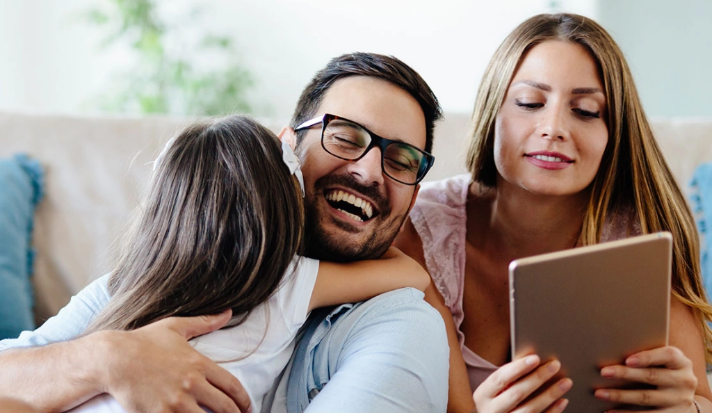 Vater und Tochter umarmen sich während Mutter auf ein Tablet schaut.