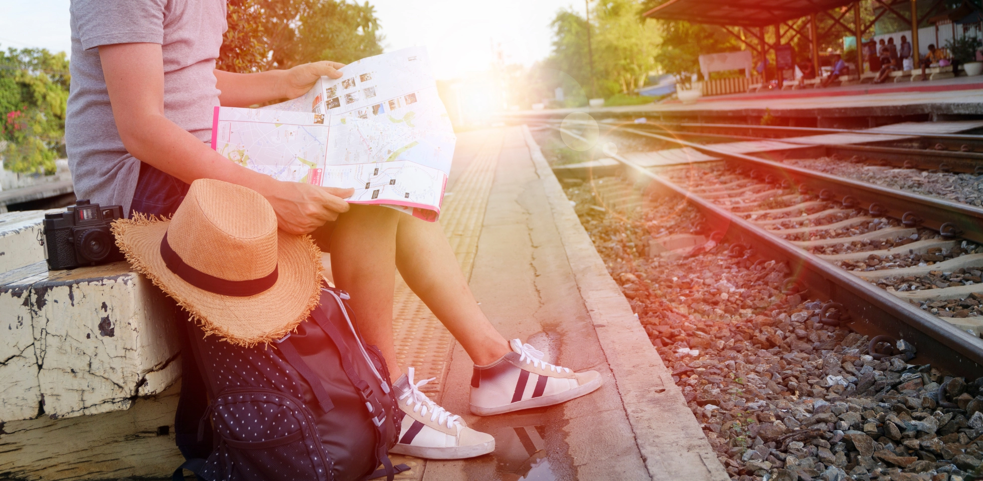 Ein Reisender sitzt wartend am Bahnhof und betrachtet eine Karte. Neben ihn steht ein kleiner Rucksack, ein Sonnenhut und eine Kamera.