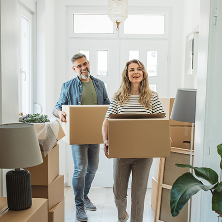 Ein Paar mittleren Alters tragen Kartons in eine helle Wohnung, in der bereits schon einige Kartons abgestellt worden sind.